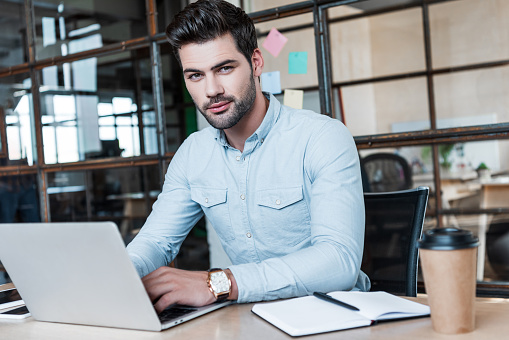 handsome young businessman using laptop and looking at camera at workplace